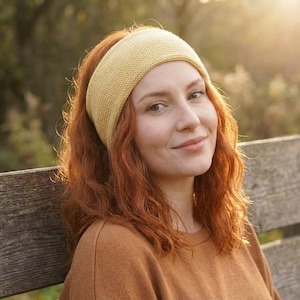 May include: A woman with red hair wearing a mustard yellow knitted headband and a brown sweater. The headband is wide and covers the forehead. The woman is smiling slightly, and the background is blurred with sunlight.