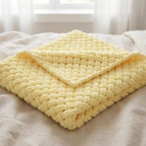 May include: A folded, square, pale yellow blanket with a woven texture. The blanket is made of a soft, plush material and is folded in a diamond shape. The blanket is resting on a neutral-coloured bedspread.
