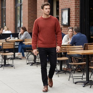 May include: A man wearing a rust-colored knit sweater and black jeans walks on a sidewalk. He is wearing brown leather boots. The background includes a brick building, outdoor seating, and other people.