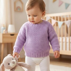 May include: A baby wearing a light purple knit sweater and white pants, holding a stuffed bunny. The sweater has a ribbed hem and cuffs. A wooden crib and other nursery items are in the background.
