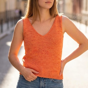 May include: Orange knitted vest top with a V-neck. The sleeveless top has a textured appearance and a ribbed hem. The model is wearing blue jeans. The background is a blurred outdoor scene.
