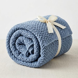 May include: Rolled-up blue knitted blanket tied with a cream-colored ribbon. The blanket has a textured pattern and is tightly rolled. The ribbon is tied in a bow, adding a decorative touch to the presentation.