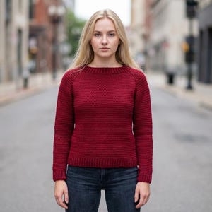 May include: A burgundy-colored sweater with a crew neck and long sleeves. The sweater is made of a knitted material and is paired with dark blue jeans. The background shows a city street.