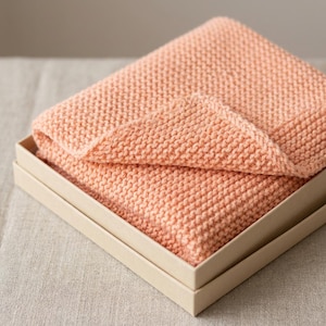 May include: A folded peach-colored knitted blanket rests inside a light beige box. The blanket is made of a thick, textured yarn, and a portion of it is folded over, revealing the knit pattern. The box is simple and rectangular.