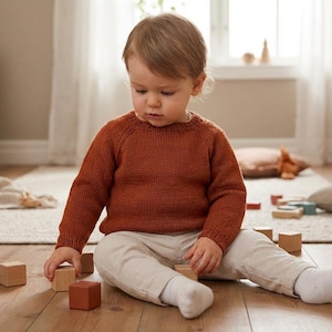 May include: A young child wearing a rust-colored sweater and light-colored pants sits on a wooden floor, surrounded by wooden blocks. The child is focused on the blocks, suggesting a playtime scene.