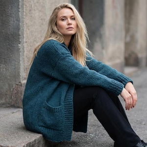 May include: A teal-colored, long-sleeved cardigan with two front pockets. The cardigan is made of a thick, knitted material. The person in the image is wearing black pants and black shoes.
