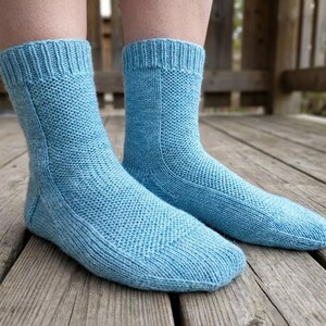 Puede incluir: Un par de calcetines de punto azul claro. Los calcetines tienen un puño acanalado y un pie texturizado. Los calcetines están hechos de un hilo suave. Los calcetines se muestran sobre una superficie de madera.