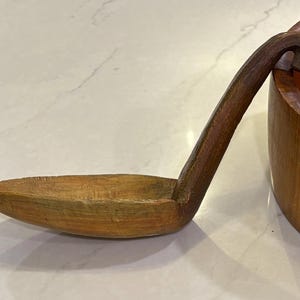 May include: A carved wooden ladle with a long, curved handle and a deep, oval-shaped bowl, positioned next to a large, round wooden bowl. The wood features a natural grain pattern with varying shades of brown, showing visible tool marks.