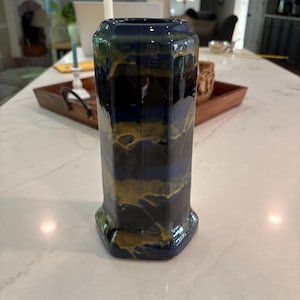 May include: A hexagonal ceramic vase featuring a glossy finish with a marbled pattern of deep navy blue and metallic gold. The vase has a straight, vertical structure with defined edges and a wide, open top, showcasing a blend of dark and light tones.