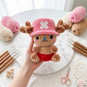 May include: A crocheted plush toy of a cartoon character, with a pink hat and white cross, brown antlers, and red shorts. The image shows the toy held in a hand, with yarn, crochet hooks, and craft supplies in the background.