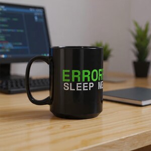 May include: Black ceramic mug with the text "ERROR SLEEP ME" in green and white. The mug is on a wooden desk, with a computer monitor and keyboard in the background. A small plant and a notebook are also visible.