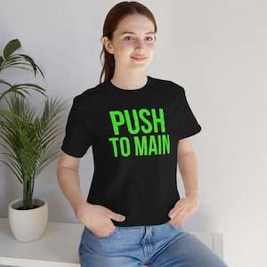 May include: Black t-shirt featuring the words "PUSH TO MAIN" in vivid green lettering. The t-shirt has a crew neck design and is likely made from a soft fabric. The model is wearing blue jeans.