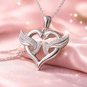 May include: A silver pendant necklace featuring a heart-shaped design with two swans and wings, embellished with small, sparkling stones. The necklace hangs on a delicate chain, set against a soft pink background.