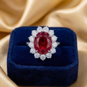 Puede incluir: Un impresionante anillo con una gran gema roja oscura de corte ovalado, rodeada por un halo de piedras blancas brillantes más pequeñas. El anillo está engarzado en una banda plateada y se presenta en una caja de terciopelo azul oscuro.