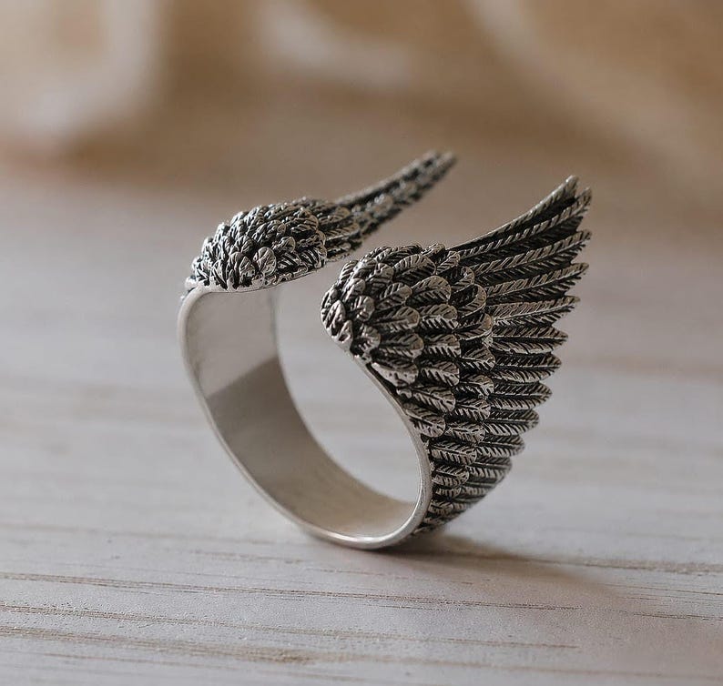 May include: A silver-colored ring with an open design, featuring detailed wing-shaped accents. The wings are textured with feather-like patterns, creating a three-dimensional effect. The ring is set against a neutral background, highlighting its intricate design.