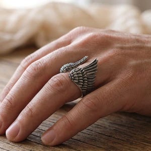 May include: A silver-toned ring featuring a detailed wing design. The ring wraps around the finger, with one wing extending from the side and another forming the top. The ring's intricate feather detailing adds a unique, artistic flair.