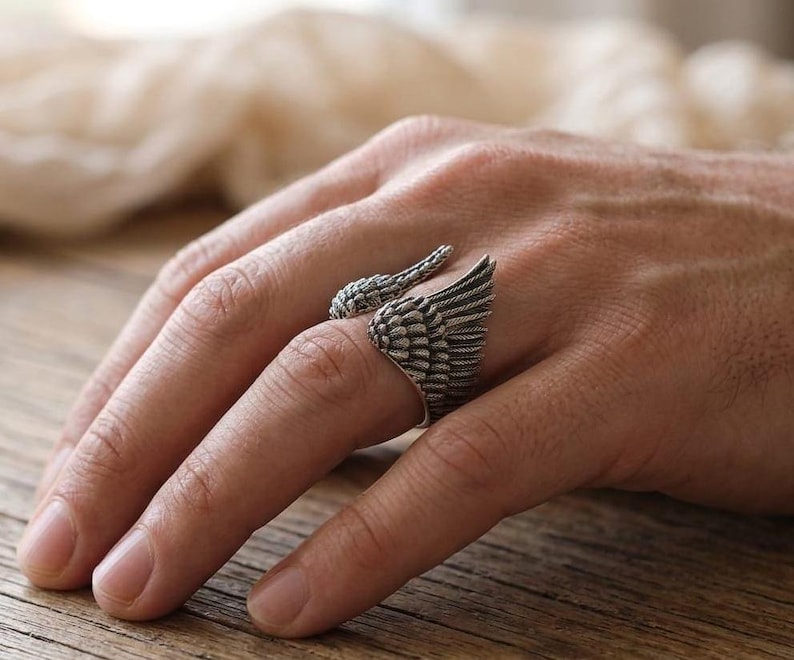 May include: A silver-toned ring featuring a detailed wing design. The ring wraps around the finger, with one wing extending from the side and another forming the top. The ring's intricate feather detailing adds a unique, artistic flair.