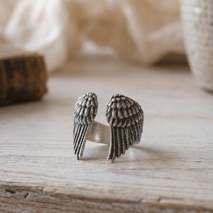 May include: A silver-colored ring featuring a detailed wing design on each side. The wings are textured with feather details, creating a realistic appearance. The ring is set against a light-colored wooden surface.