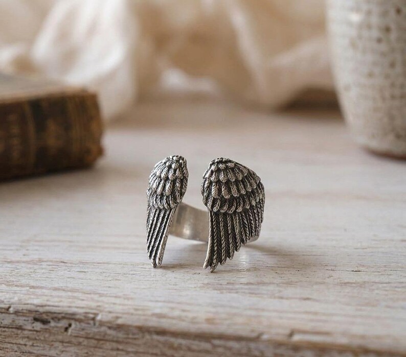 May include: A silver-colored ring featuring a detailed wing design on each side. The wings are textured with feather details, creating a realistic appearance. The ring is set against a light-colored wooden surface.
