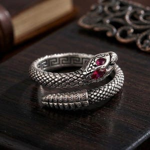 Può includere: Un anello a forma di serpente argentato con occhi di gemme rosse. L'anello è a spirale, con la testa del serpente sollevata e la coda che forma l'altra estremità. L'anello ha un motivo strutturato a scaglie e un motivo a chiave greca.