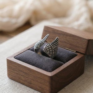 May include: A silver-colored ring shaped like angel wings, displayed in a wooden jewelry box with a dark gray velvet interior. The ring's intricate feather details are visible. The box is open, revealing the ring's design.