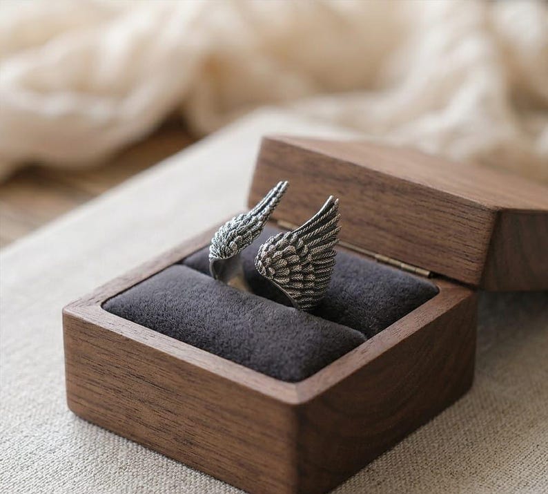 May include: A silver-colored ring shaped like angel wings, displayed in a wooden jewelry box with a dark gray velvet interior. The ring's intricate feather details are visible. The box is open, revealing the ring's design.