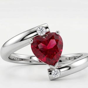 May include: A silver ring with a heart-shaped, deep red gemstone at the center. Two smaller, round, clear gemstones are set on either side of the heart. The ring's band curves around the heart-shaped stone.