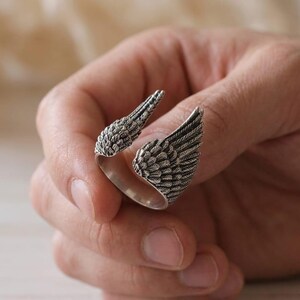 May include: A silver-colored ring with an open design, featuring two detailed wing shapes. The wings are textured with feather-like patterns, creating a realistic appearance. The ring is designed to wrap around the finger.