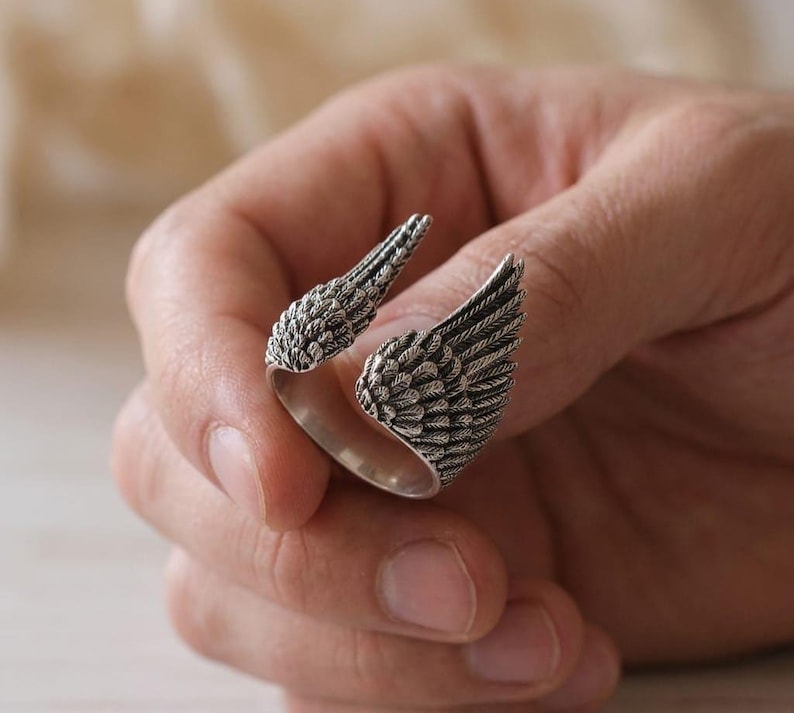 May include: A silver-colored ring with an open design, featuring two detailed wing shapes. The wings are textured with feather-like patterns, creating a realistic appearance. The ring is designed to wrap around the finger.