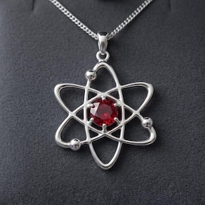 May include: A silver pendant necklace with an atom-shaped design. The pendant has a central, round, red gemstone and is suspended from a fine chain. The necklace is presented against a dark grey background.