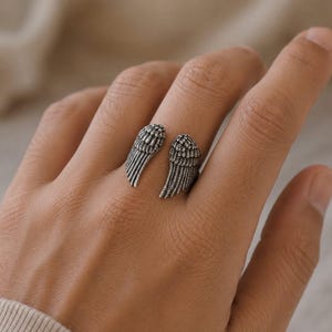 May include: A silver-toned ring featuring an open design with two detailed wing shapes. The wings are positioned to wrap around the finger, creating a unique and symbolic accessory. The ring's design showcases intricate feather detailing.