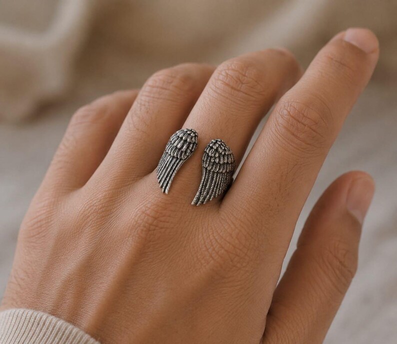 May include: A silver-toned ring featuring an open design with two detailed wing shapes. The wings are positioned to wrap around the finger, creating a unique and symbolic accessory. The ring's design showcases intricate feather detailing.