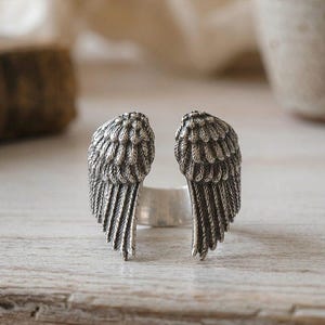 May include: A silver-toned ring featuring a detailed design of two open wings. The wings are textured with feather details, creating a realistic appearance. The ring is set against a light-colored wooden surface.