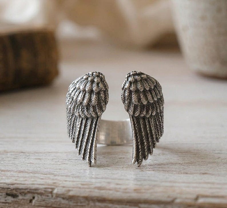 May include: A silver-toned ring featuring a detailed design of two open wings. The wings are textured with feather details, creating a realistic appearance. The ring is set against a light-colored wooden surface.