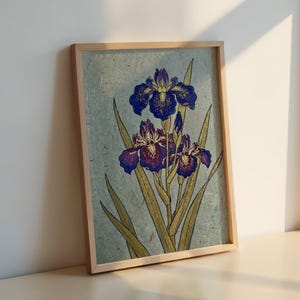 May include: A framed botanical print featuring deep purple iris flowers with yellow accents and long green leaves. The artwork is set against a textured, light blue-grey background, enclosed in a simple, light-toned wooden frame.