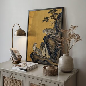 A traditional East Asian sumi-e style painting featuring two snow leopards on rocky cliffs beside a flowing waterfall, with a twisted pine branch and white plum blossoms cascading from above, set against a warm luminous gold leaf background with small birds in the distance in muted earthy tones.