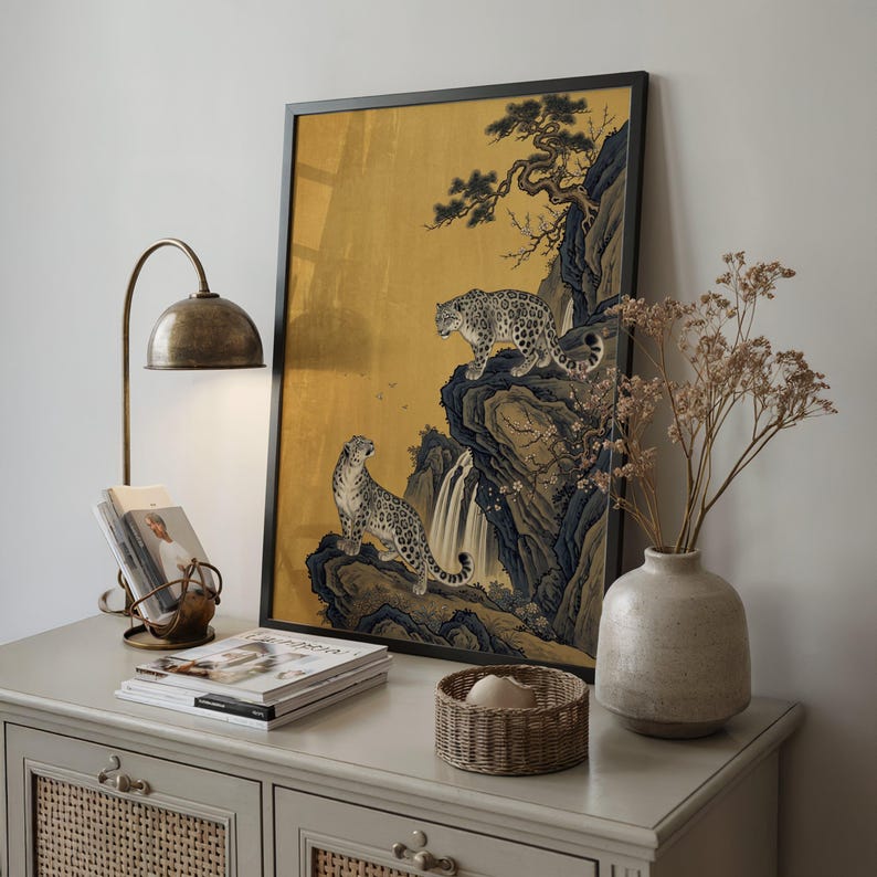 A traditional East Asian sumi-e style painting featuring two snow leopards on rocky cliffs beside a flowing waterfall, with a twisted pine branch and white plum blossoms cascading from above, set against a warm luminous gold leaf background with small birds in the distance in muted earthy tones.