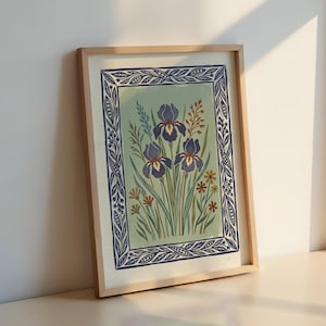 May include: Framed floral art print featuring three purple iris flowers with yellow centers, surrounded by green foliage and small orange and yellow flowers. The artwork is set against a light green background and framed in a light wood frame with a blue border.