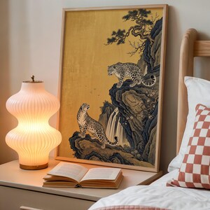 A traditional East Asian sumi-e style painting featuring two snow leopards on rocky cliffs beside a flowing waterfall, with a twisted pine branch and white plum blossoms cascading from above, set against a warm luminous gold leaf background with small birds in the distance in muted earthy tones.