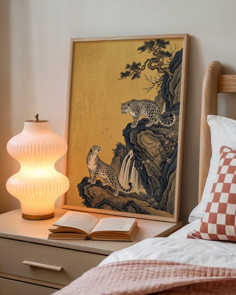 A traditional East Asian sumi-e style painting featuring two snow leopards on rocky cliffs beside a flowing waterfall, with a twisted pine branch and white plum blossoms cascading from above, set against a warm luminous gold leaf background with small birds in the distance in muted earthy tones.