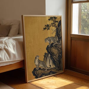A traditional East Asian sumi-e style painting featuring two snow leopards on rocky cliffs beside a flowing waterfall, with a twisted pine branch and white plum blossoms cascading from above, set against a warm luminous gold leaf background with small birds in the distance in muted earthy tones.