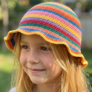 May include: A crochet bucket hat featuring horizontal stripes in vibrant colors including orange, pink, purple, green, blue, and red. The hat has a rounded crown and a wavy, yellow-trimmed brim, crafted with a dense, textured yarn stitch pattern.