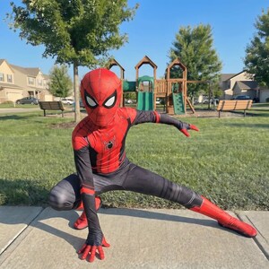 May include: A red and black Spider-Man costume with a mask, worn by a person crouching on a sidewalk. The costume features a spider emblem on the chest and red gloves and boots. The background includes a playground and green grass.