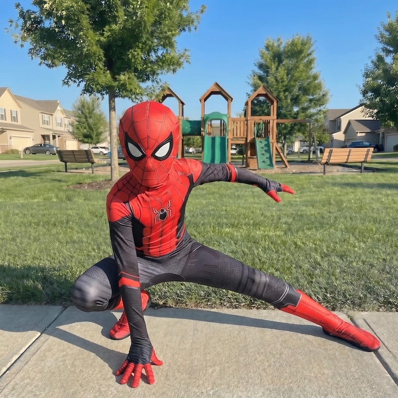 May include: A red and black Spider-Man costume with a mask, worn by a person crouching on a sidewalk. The costume features a spider emblem on the chest and red gloves and boots. The background includes a playground and green grass.