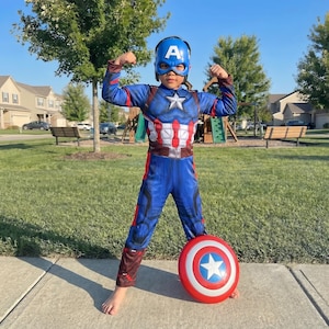 May include: A child wearing a Captain America costume, flexing their arms. The blue and red costume includes a mask, a star on the chest, and a matching shield. The shield is red, white, and blue with a white star.