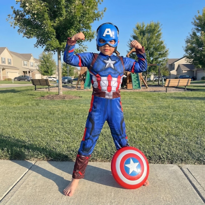 May include: A child wearing a Captain America costume, flexing their arms. The blue and red costume includes a mask, a star on the chest, and a matching shield. The shield is red, white, and blue with a white star.