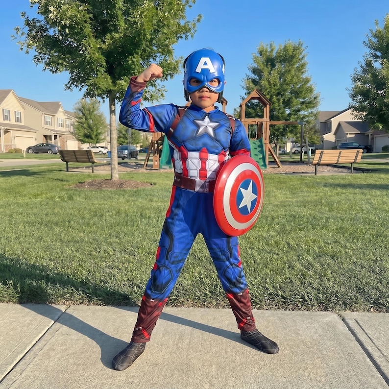 May include: A child in a Captain America costume, including a blue helmet with a white "A", a blue and red jumpsuit, and a red, white, and blue shield. The child is flexing their arm.