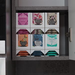 Könnte beinhalten: Ein weißer Teebeutel-Organizer mit verschiedenen Teeschachteln, darunter Pukka Morning Berry, Clipper Skinni Vanilli, English Breakfast, Mango & Citrus, Camomile Lemon & Lavender und Clipper Organic Teebeutel, angeordnet in drei Spalten.