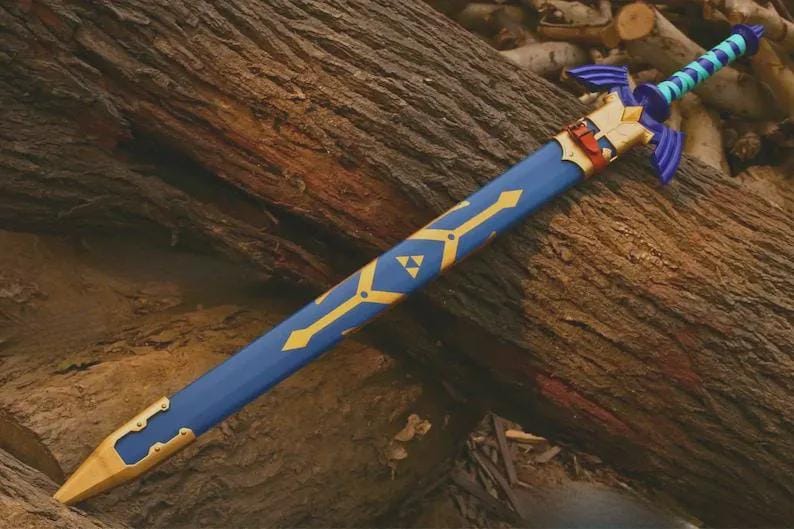 Puede incluir: Una espada azul en una vaina azul a juego con detalles dorados. La vaina presenta un s&iacute;mbolo de trifuerza dorado y una punta de metal dorado. La empu&ntilde;adura es azul con una guarda morada y un dise&ntilde;o de agarre en espiral azul claro y oscuro.
