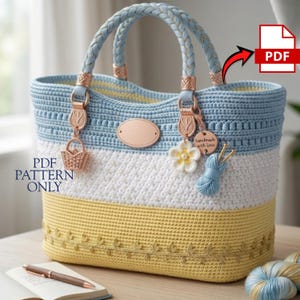 May include: A handmade crochet tote bag in blue, white, and yellow. The bag features braided blue handles, decorative charms, and a floral embellishment. The text "PDF PATTERN ONLY" is visible on the bag.